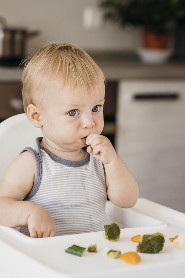 Bébé aux yeux bleus découvrant des légumes par lui-même selon l'approche DME en crèche, assis dans sa chaise haute avec brocolis et carottes