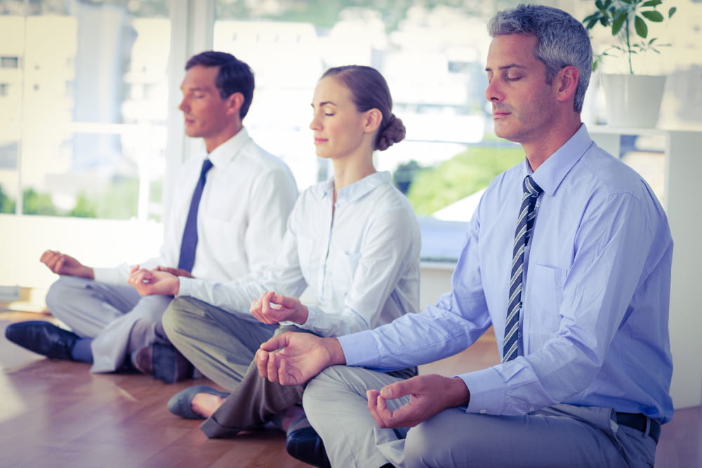 Trois collaborateurs en tenue de bureau pratiquent la méditation en groupe, illustrant les solutions de bien-être en entreprise axées sur la santé mentale.