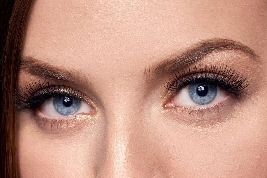 Un gros plan de deux yeux bleus intenses, maquillés avec des faux cils et un fard doré pour les sublimer.
