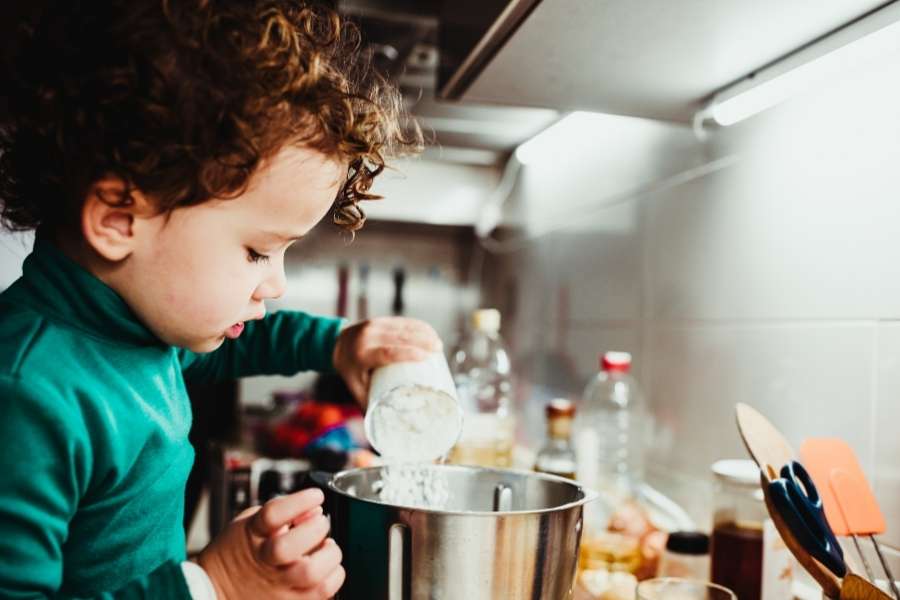 Un jeune enfant aux cheveux bouclés verse de la farine dans un bol, illustrant l'importance d'inclure des ingrédients sains dans les menus pour un toddler de deux ans.